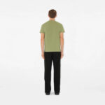 Uomo di spalle in outfit casual verde oliva e pantaloni neri su sfondo bianco minimalista.