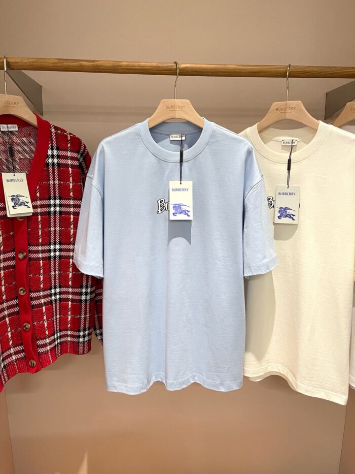 Tre magliette casual in rosso quadri, azzurro e bianco su appendini di legno.