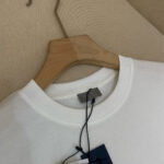 Maglietta bianca casual in cotone con tag blu, su gancio di legno naturale, sfondo beige neutro.