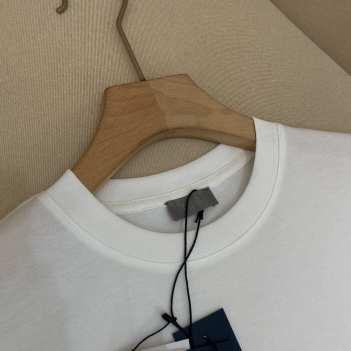 Maglietta bianca casual in cotone con tag blu, su gancio di legno naturale, sfondo beige neutro.