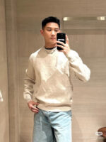 Ragazzo giovane in selfie casual in ambiente moderno, con sweater beige e jeans blu chiari.