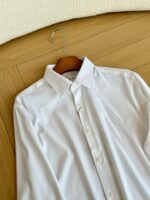 Camicia bianca classica su gancio di legno, fondo in legno chiaro, stile elegante e versatile.