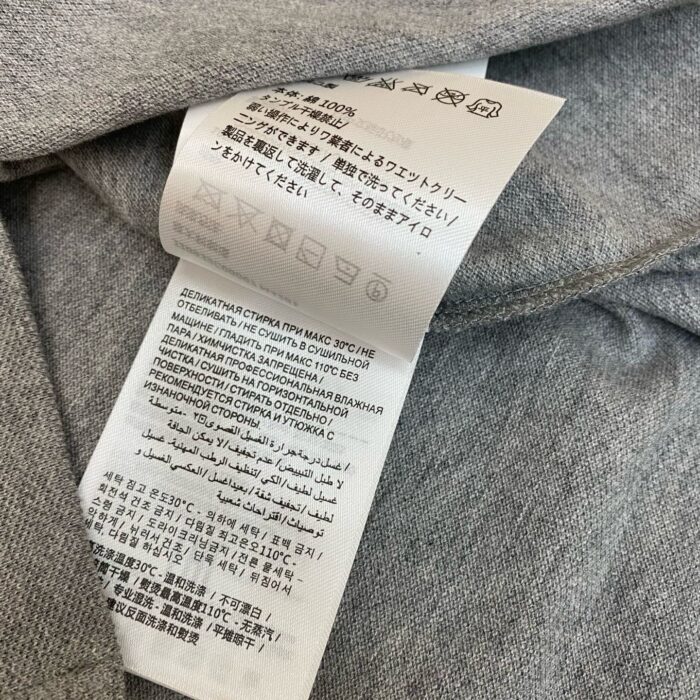 Etichetta di cura del capo con icone e istruzioni multilingua su tessuto grigio.