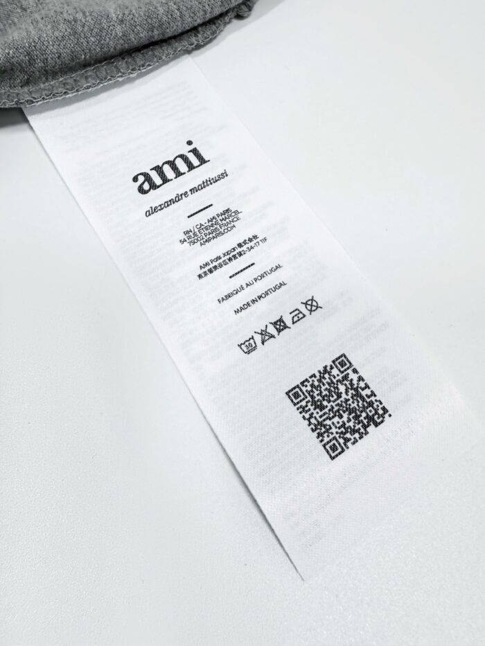 Etichetta del brand Ami con QR code e simboli di cura su tessuto grigio.