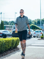 Uomo in stile casual con tatuaggi e occhiali, passeggia in area ricreativa estiva.