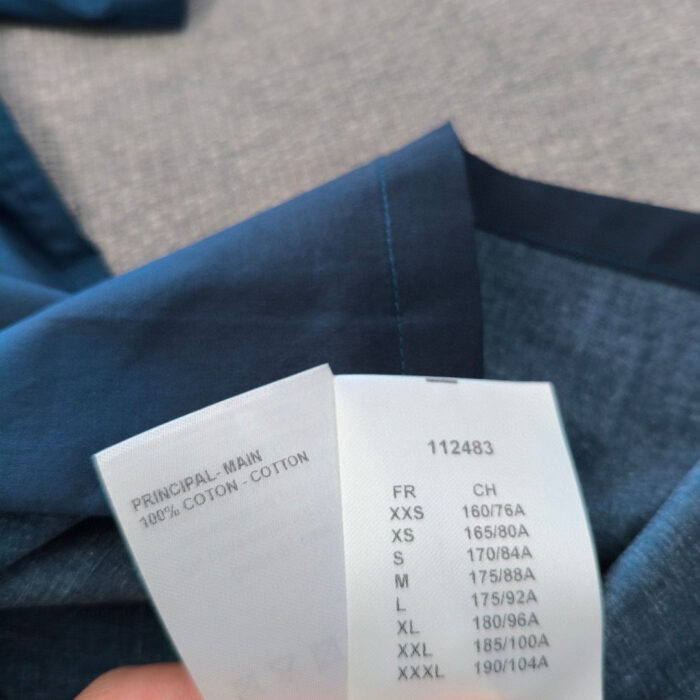 Dettaglio della etichetta e della camicia in cotone blu con informazioni di taglia e cura.