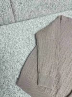 Maglione beige a maglia con motivo a trecce su sfondo grigio accogliente e testurizzato.