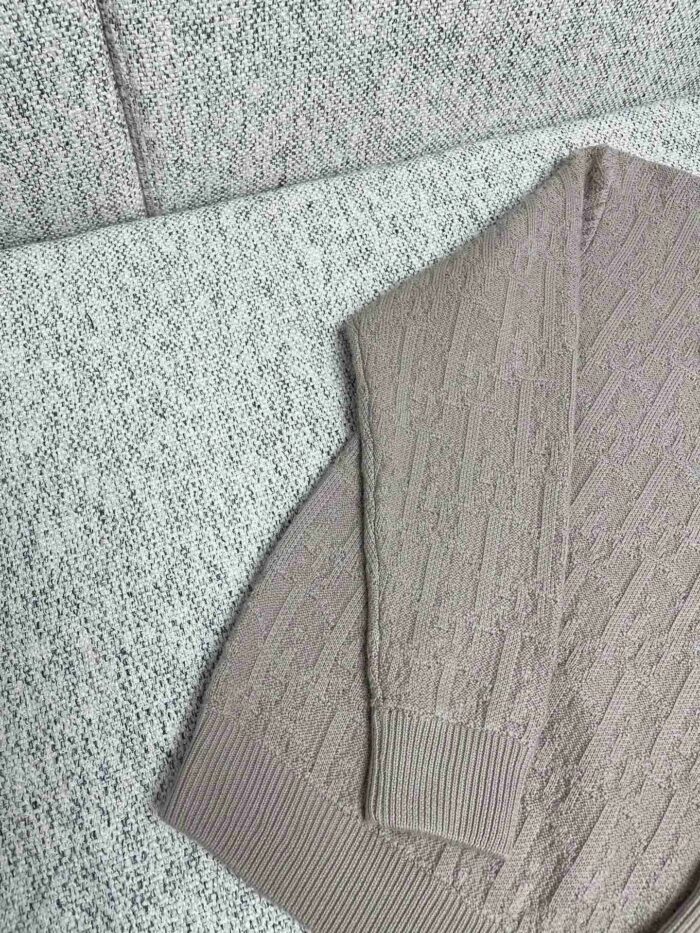Maglione beige a maglia con motivo a trecce su sfondo grigio accogliente e testurizzato.