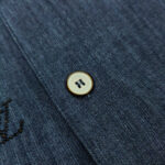 Dettaglio di camicia in denim, bottoni e ricamo stilizzato, stile casual e qualità.