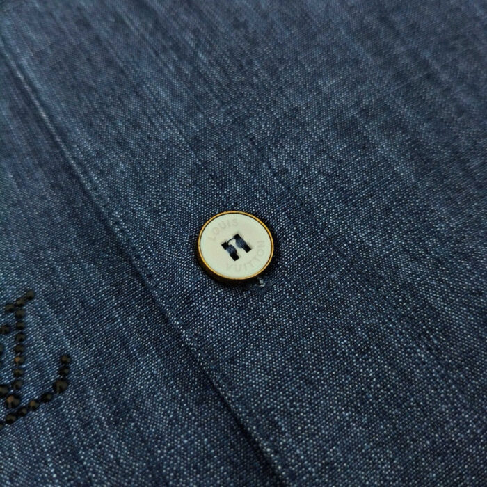 Dettaglio di camicia in denim, bottoni e ricamo stilizzato, stile casual e qualità.