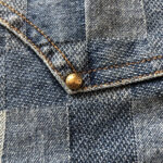Dettaglio denim plaid con rivetto in ottone e cuciture beige, stile vintage e qualità artigianale.