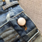 Dettaglio di Jeans con bottone metallico e cuciture contrasto su denim usurato.