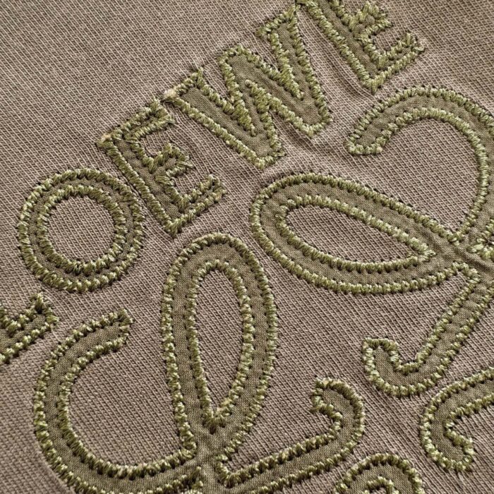 Ricamo LOEWE su tessuto neutro con dettagli eleganti e artigianali in verde oliva.