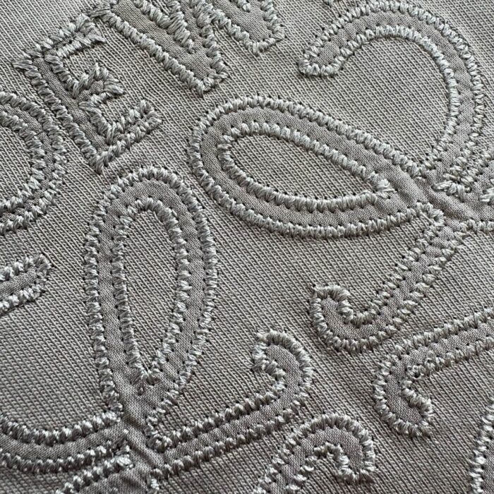 Ricamo elegante su tessuto neutro con lettere e motivi swirl, ideale per decorazioni raffinate.