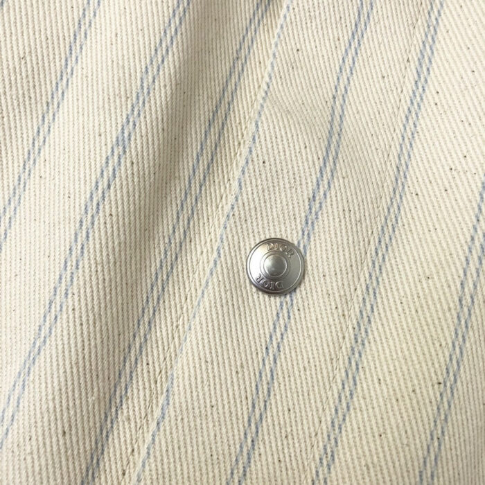 Tessuto beige con righe blu e bottone metallico elegante, ideale per moda e arredamento.