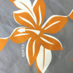 Design floreale elegante con logo Gucci su sfondo grigio, floreali arancioni e foglie verdi.