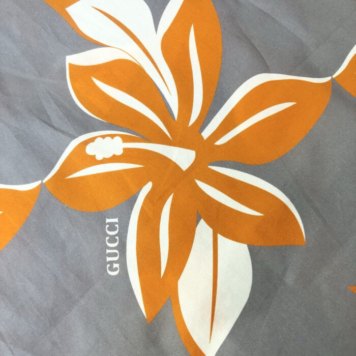 Design floreale elegante con logo Gucci su sfondo grigio, floreali arancioni e foglie verdi.
