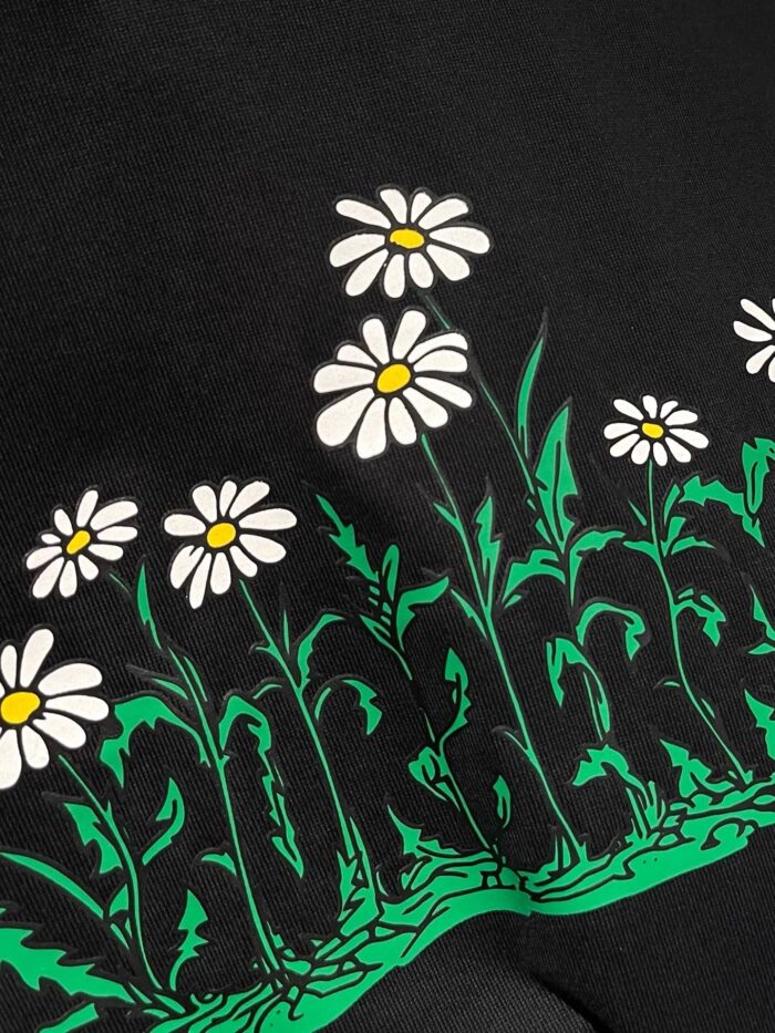 Maglietta con fiori di margherita e testo GARDEN su sfondo nero.
