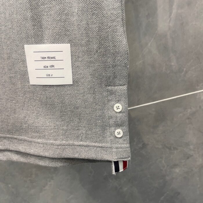 Camicia grigia con bottoni bianchi e dettaglio a righe rosse, bianche e blu sullorlo.