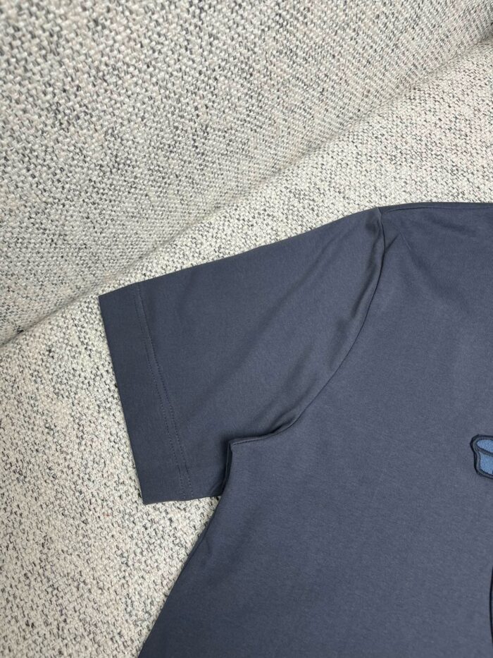 Dettaglio manica di una t-shirt grigia casual su sfondo tessuto neutro.