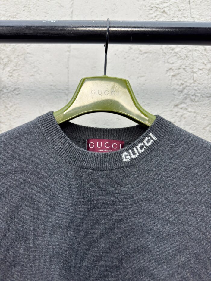 Maglione grigio Gucci con logo sul collo, su hanger verde elegante e sfondo testurizzato.
