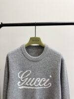 Maglione grigio Gucci con logo ricamato, stile elegante e casual, su appendiabiti di alta qualità.