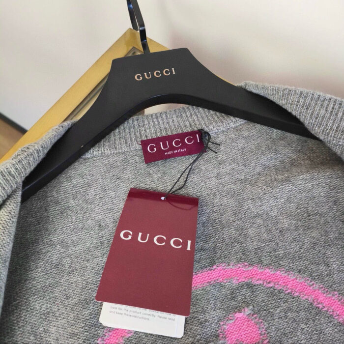 Maglione Gucci grigio con Logo rosa, etichetta marrone, su appendiabiti di legno elegante.