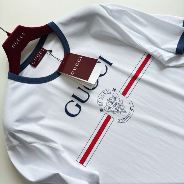 T-shirt bianco Gucci con logo blu, dettagli crest e strisce rosse sportive.