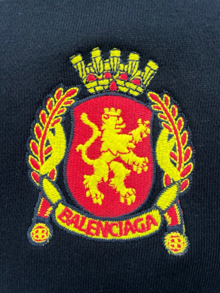 Emblema Balenciaga con leone, corona, lauro e dettagli dorati su sfondo scuro.