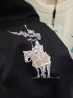 Ricamo di cavaliere con bandiera su sfondo nero, simbolo di forza e eleganza sartoriale.