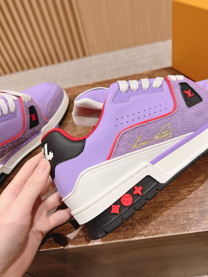 Sneakers lavanda con dettagli rossi, nero opaco e logo dorato su superficie in legno.
