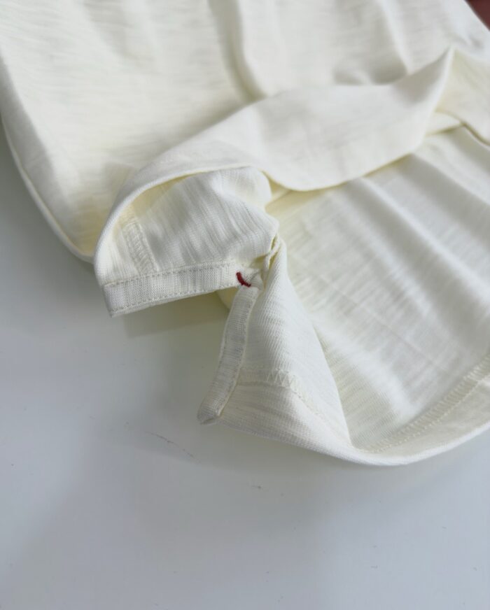 Dettaglio di camicia in lino crema con cuciture precise e piccolo dettaglio rosso.