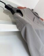 Camicia grigia casual elegante con tasca sul petto, appesa a gancio nero su sfondo bianco minimal.