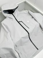 Giacca leggera waterproof bianca con cappuccio, tasca frontale nera e design minimalista elegante.