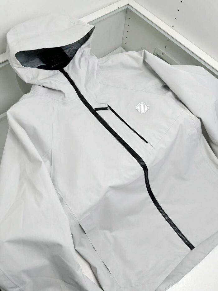 Giacca leggera waterproof bianca con cappuccio, tasca frontale nera e design minimalista elegante.