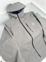 Giacca leggera grigia con cappuccio e tasche zip, stile minimalista e protezione outdoor.