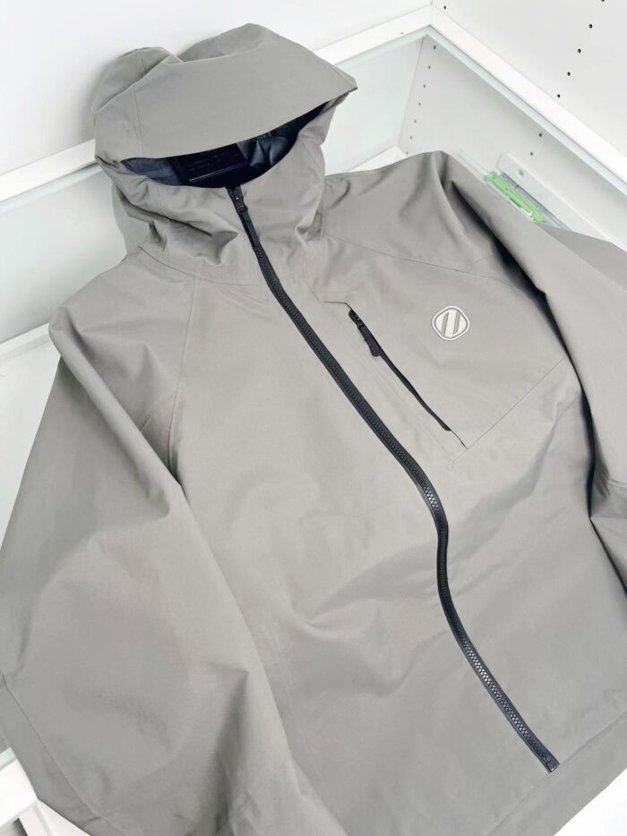 Giacca leggera grigia con cappuccio e tasche zip, stile minimalista e protezione outdoor.
