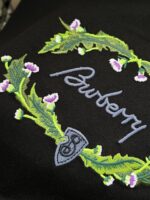 Ricamo elegante con testo Burberry, foglie, fiori lavanda e scudo simbolo di tradizione.