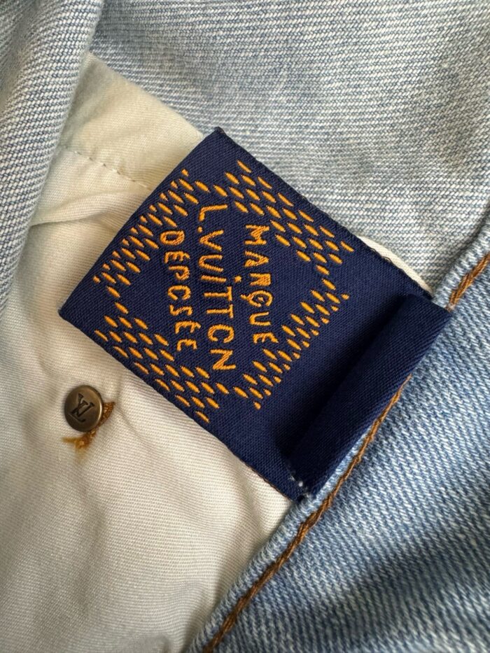 Etichetta Louis Vuitton blu con logo dorato su denim chiaro e bottone metallico elegante.