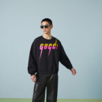 Modello con felpa Gucci, pantaloni in pelle e occhiali da sole trendy, stile casual elegante.