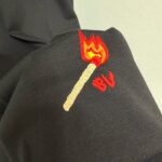 Ricamo di fiammifero con fiamma e lettere BV su tessuto scuro.