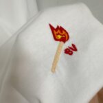 Ricamo di fiammifero con fiamma colorata su maglietta bianca, dettaglio creativo e moderno.