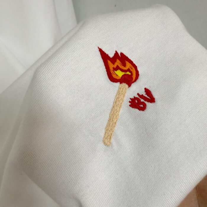 Ricamo di fiammifero con fiamma colorata su maglietta bianca, dettaglio creativo e moderno.