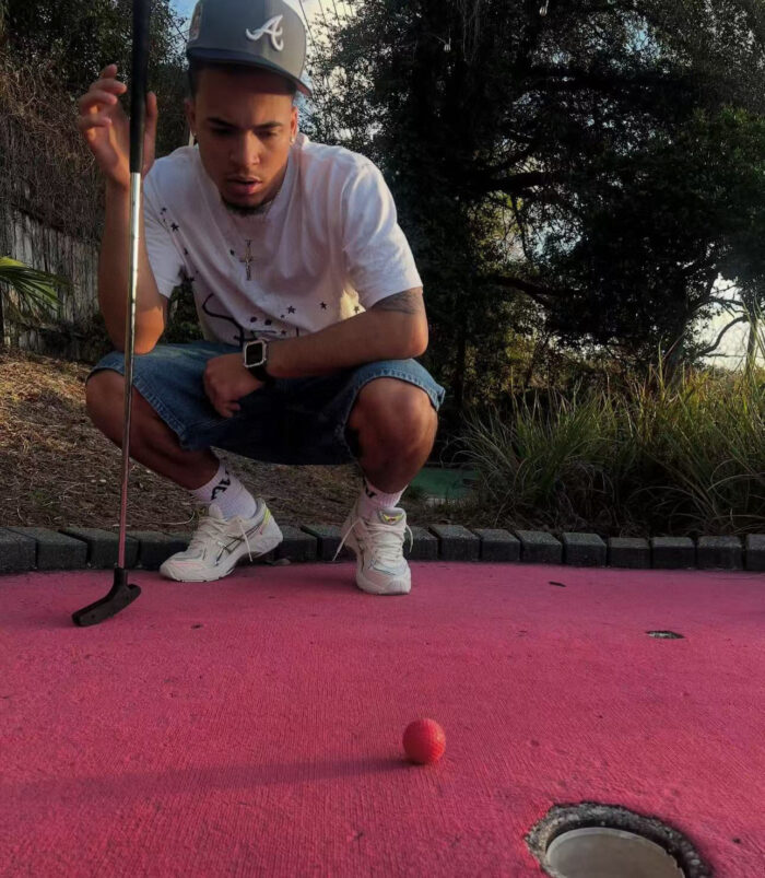 Giovane che gioca a mini golf allaperto su green rosa con natura e atmosfera estiva.