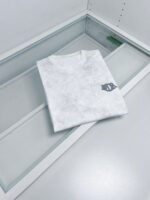 Maglietta bianca minimalista su vetrina moderna, logo in alto a sinistra, ambiente luminoso e raffinato.