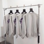 Magliette grigie con scritte viola, stile streetwear, display moderno su scaffale metallico.
