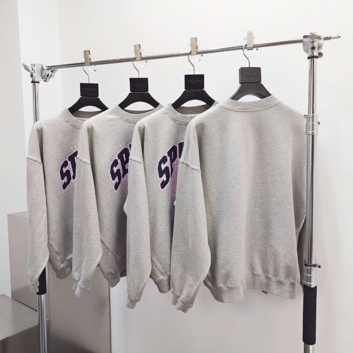 Magliette grigie con scritte viola, stile streetwear, display moderno su scaffale metallico.