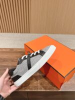 Sneaker grigia con dettagli neri, lacci bianchi, scatola arancione su sfondo in legno elegante.