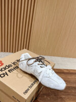 Sneaker bianca moderna su scatola in cartone con logo New Balance e sfondo minimalista in legno.