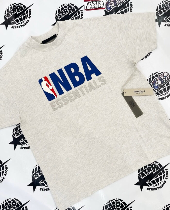 Maglietta grigia NBA Essentials in stile streetwear, con logo e scritte blu vivaci.